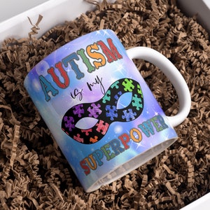 Puede incluir: Una taza de cer&aacute;mica blanca con un dise&ntilde;o de galaxia azul, morado y rosa. La taza tiene el texto "El autismo es mi superpoder" y una m&aacute;scara de superh&eacute;roe negra con piezas de rompecabezas.