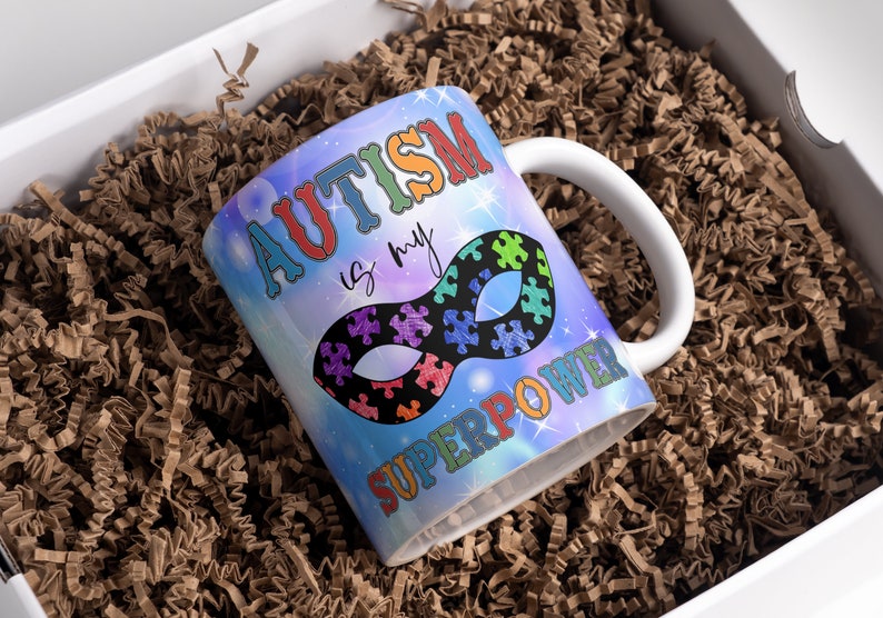 Puede incluir: Una taza de cer&aacute;mica blanca con un dise&ntilde;o de galaxia azul, morado y rosa. La taza tiene el texto "El autismo es mi superpoder" y una m&aacute;scara de superh&eacute;roe negra con piezas de rompecabezas.