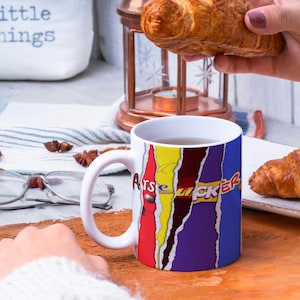May include: White ceramic mug with a colorful design featuring the words "ARSE LICKER" in a comic book style. The mug is filled with a brown liquid and is sitting on a wooden surface. There are two croissants in the background.