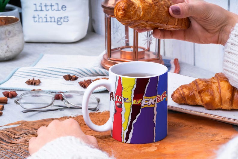May include: White ceramic mug with a colorful design featuring the words "ARSE LICKER" in a comic book style. The mug is filled with a brown liquid and is sitting on a wooden surface. There are two croissants in the background.