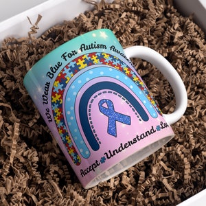 Puede incluir: Una taza de cer&aacute;mica blanca con un dise&ntilde;o de arco&iacute;ris y el texto "We wear blue for autism awareness accept understand love". El arco&iacute;ris est&aacute; hecho de piezas de rompecabezas y una cinta azul est&aacute; en el centro.