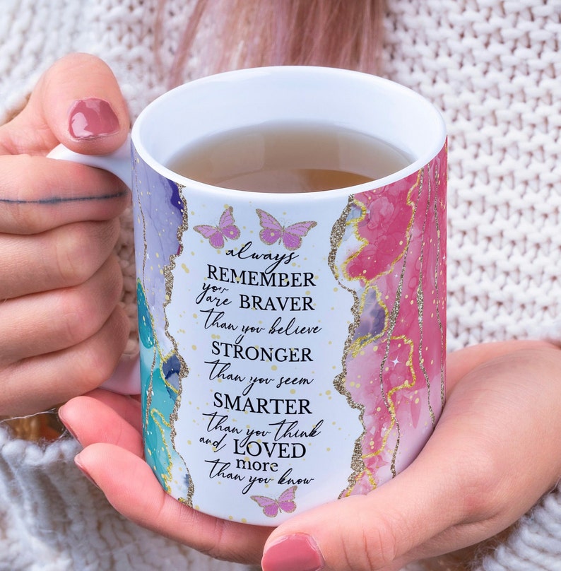 May include: White ceramic mug with a pink, blue, and green abstract design. The mug has gold glitter accents and butterflies. The text on the mug reads "always remember you are braver than you believe stronger than you seem smarter than you think and loved more than you know."