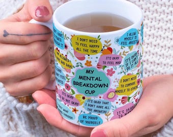 May include: A white ceramic mug with the text "My Mental Breakdown Cup" and various affirmations in a colorful design. The mug is held by a person with pink nail polish.
