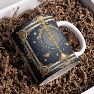 May include: A white ceramic mug with a blue and gold design featuring a zodiac scale symbol. The mug is in a white box with brown packing paper.