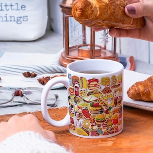May include: White ceramic mug with a colorful design of various fast food items, such as burgers, fries, pizza, and donuts. The mug is filled with a brown liquid and is sitting on a wooden surface. A hand is holding a croissant above the mug.
