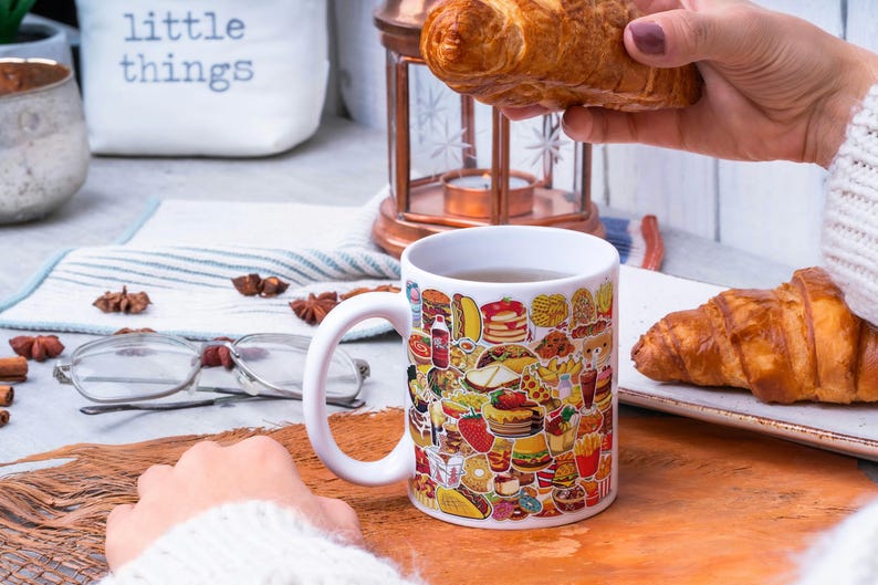 May include: White ceramic mug with a colorful design of various fast food items, such as burgers, fries, pizza, and donuts. The mug is filled with a brown liquid and is sitting on a wooden surface. A hand is holding a croissant above the mug.