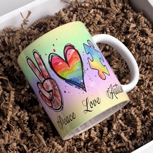 Puede incluir: Una taza de cer&aacute;mica blanca con un dise&ntilde;o colorido que presenta un signo de paz, un coraz&oacute;n arco&iacute;ris y una pieza de rompecabezas. El texto "Peace Love Autism" est&aacute; escrito en la taza.