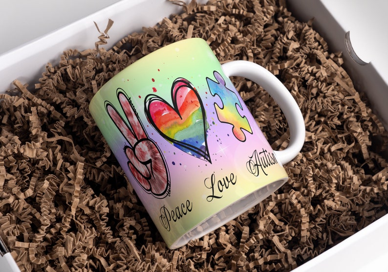 Puede incluir: Una taza de cer&aacute;mica blanca con un dise&ntilde;o colorido que presenta un signo de paz, un coraz&oacute;n arco&iacute;ris y una pieza de rompecabezas. El texto "Peace Love Autism" est&aacute; escrito en la taza.