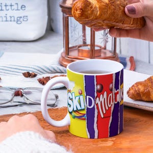 May include: White ceramic mug with a colorful design featuring the words "Skid  Marks" and a chocolate bar. The mug is filled with a brown liquid and is sitting on a wooden surface. There are two croissants in the background.