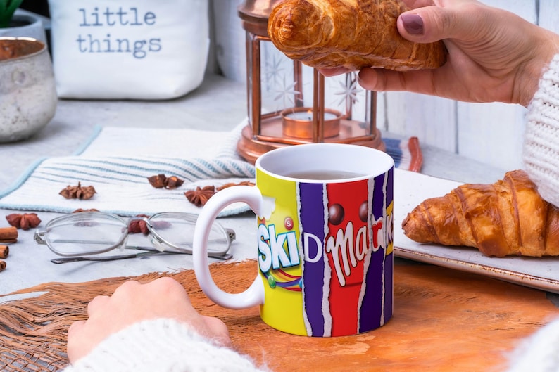 May include: White ceramic mug with a colorful design featuring the words "Skid  Marks" and a chocolate bar. The mug is filled with a brown liquid and is sitting on a wooden surface. There are two croissants in the background.