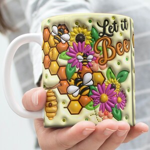 Könnte beinhalten: Weiße Keramiktasse mit einem gelben, braunen und violetten Bienendesign. Die Tasse hat einen Henkel und trägt den Schriftzug "Let it Bee" in schwarzer Schrift.
