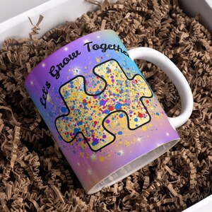 Puede incluir: Una taza de cer&aacute;mica blanca con un fondo degradado morado, azul y amarillo. La taza presenta una gran pieza de rompecabezas amarilla con salpicaduras de pintura de colores y el texto "Let's Grow Together".