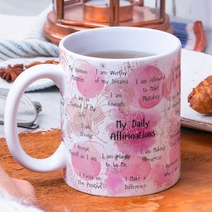 May include: White ceramic mug with a pink and gold design featuring watercolor-style flowers and the text "My Daily Affirmations". The text includes positive affirmations such as "I am worthy of my dreams", "I am enough", and "I am happy to be me".