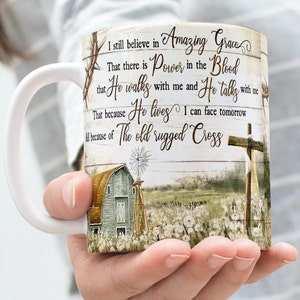 May include: White ceramic mug with a rustic design featuring a barn, windmill, and a cross. The mug has a brown handle and a quote that reads: "I still believe in Amazing Grace. That there is Power in the Blood that He walks with me and He talks with me. That because He lives I can face tomorrow. All because of The old rugged Cross."