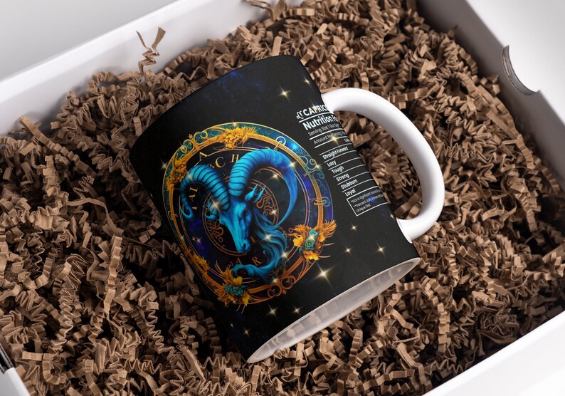 May include: A white ceramic mug with a black and gold design featuring a blue ram's head with horns. The mug has a black background with gold stars and a gold circle around the ram's head. The text "CAPRICORN" is printed on the mug, along with a list of "Nutritional Facts" for the zodiac sign.