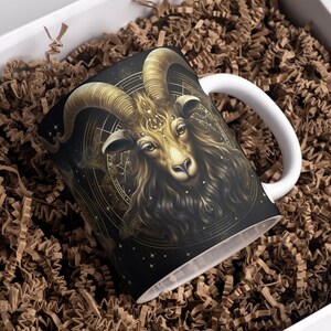 May include: A white ceramic mug with a black and gold design featuring a ram's head with intricate details. The ram's head is surrounded by a circular design with stars and other symbols.