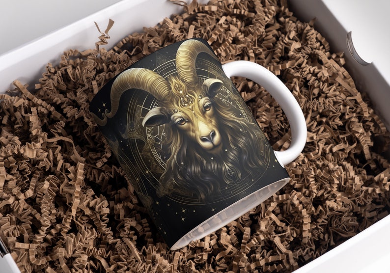 May include: A white ceramic mug with a black and gold design featuring a ram's head with intricate details. The ram's head is surrounded by a circular design with stars and other symbols.