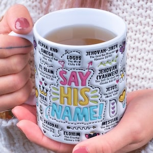 May include: White ceramic mug with a colorful design featuring the names of God in different languages, including "Say His Name!" in large, bold letters. The mug is filled with a light brown liquid.