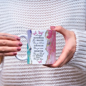 May include: White ceramic mug with a pink, green, and blue abstract design. The mug has a gold rim and the text "always remember you are braver than you believe stronger than you seem smarter than you think and loved more than you know" printed on it.