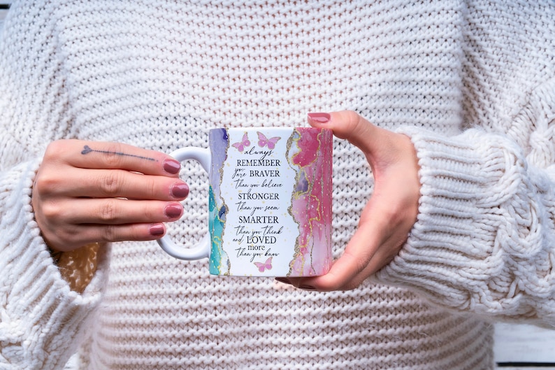 May include: White ceramic mug with a pink, green, and blue abstract design. The mug has a gold rim and the text "always remember you are braver than you believe stronger than you seem smarter than you think and loved more than you know" printed on it.