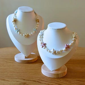 Può includere: Due collane esposte su busti bianchi, caratterizzate da perle bianche alternate a piccole stelle marine e ciondoli a forma di conchiglia in tonalità pastello come azzurro, beige, rosa e bianco, disposti in un motivo lungo i fili.