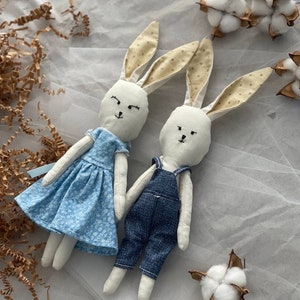 Puede incluir: Dos juguetes de peluche de conejo hechos a mano. Un conejo lleva un vestido floral azul y blanco y el otro lleva un mono azul. Ambos conejos tienen pelaje blanco y ojos negros.
