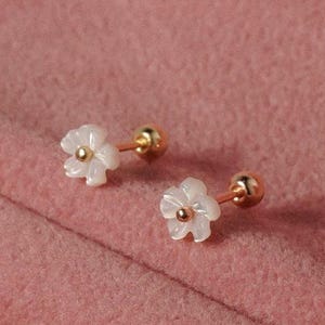 Puede incluir: Un par de pendientes de botón con forma de flor. Cada pendiente presenta un pequeño diseño en forma de flor blanca con un centro dorado y un poste dorado. Los pendientes se muestran sobre una superficie rosa texturizada.
