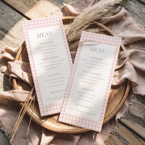 Op de afbeelding: Twee menu's met een roze gingham rand, met een menu voor een maaltijd. De menu's liggen op een houten dienblad, met gedroogde planten en een roze doek. Het menu bevat voorgerechten, hoofdgerechten, desserts en drankjes.