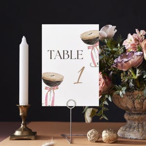 May include: White card with the text "TABLE 1" in brown and black. The card has two illustrations of coffee drinks with pink ribbons. The card is on a metal stand.