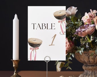 May include: White card with the text "TABLE 1" in brown and black. The card has two illustrations of coffee drinks with pink ribbons. The card is on a metal stand.