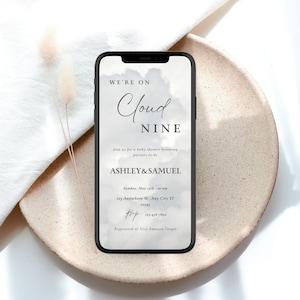 May include: A smartphone displays a baby shower invitation with the text "WE'RE ON Cloud NINE." The invitation includes the names Ashley & Samuel, date, time, and RSVP information. The phone rests on a beige plate with a white cloth and dried flowers.
