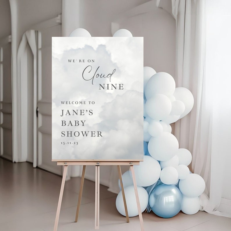 May include: A baby shower sign with a blue and white cloudy background. The sign reads "We're on Cloud Nine. Welcome to Jane's Baby Shower 15.11.23".