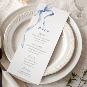 May include: A white menu card featuring a blue watercolour bow design, resting on a white plate. The menu lists starters, main dishes, desserts, and beverages. Menu items include smoked salmon, grilled rack of lamb, and white chocolate mousse.