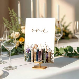 Può includere: Un segnaposto bianco con la parola "one" in un carattere dorato. La parte inferiore della carta presenta un'illustrazione ad acquerello di libri e fiori di campo. La carta è tenuta in un supporto di metallo dorato, con una tovaglia bianca sullo sfondo.