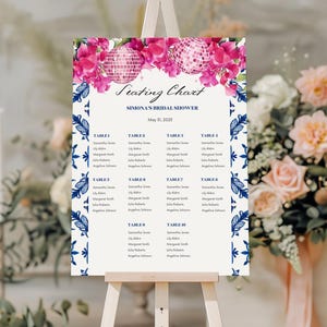 May include: A seating chart for a bridal shower, displayed on an easel. The design features a floral and disco ball motif in pink and white, with a blue and white border. The text reads "Seating Chart" and "Simona's Bridal Shower".