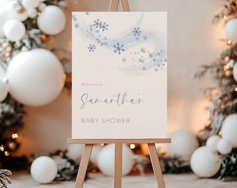 Puede incluir: Un letrero beige claro sobre un caballete de madera, con detalles de copos de nieve azules y las palabras "Welcome to Samantha's Baby Shower" en una elegante escritura. El letrero está rodeado de globos blancos y decoraciones festivas.