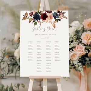 May include: A seating chart for Emily's bridal shower, displayed on an easel. The chart features a floral design with burgundy, navy, and blush tones. The text includes the date, table numbers, and guest names.