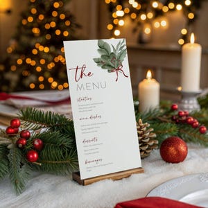 May include: A white menu card with the text "The MENU" in red script, featuring a watercolor illustration of green leaves and red berries. The menu is displayed on a wooden stand, with a festive holiday theme.