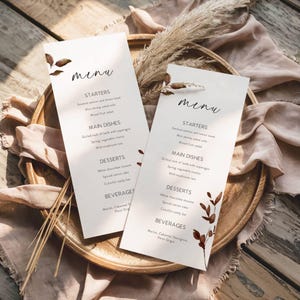Peut inclure: Deux élégants menus avec le mot "menu" en écriture stylisée en haut. Les cartes présentent une liste d'entrées, de plats principaux, de desserts et de boissons. Des accents floraux bruns et du texte sont imprimés sur un fond crème. Les menus sont sur un plateau en bois.