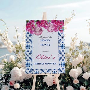 May include: A sign on a wooden easel, featuring pink floral and disco ball designs. The sign displays the text "She found her HONEY HONEY Welcome to Chloe's BRIDAL SHOWER". The background includes white flowers and foliage.