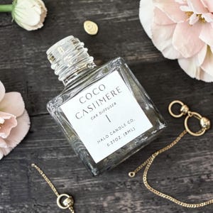 May include: A square glass car diffuser bottle with a white label reading COCO CASHMERE CAR DIFFUSER HALO CANDLE CO. 0.27OZ. (8ML). The bottle has a clear threaded neck and is positioned near gold-toned metal chains and light pink flowers.