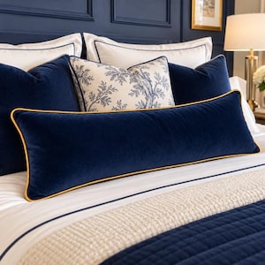 May include: A long, rectangular navy blue velvet lumbar pillow with gold piping trim, positioned in front of two square navy blue velvet pillows and a central decorative pillow featuring a blue and white botanical pattern on a bed.