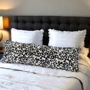 Peut inclure: Un lit blanc avec un coussin de traversin à imprimé léopard noir et blanc sur un couvre-lit blanc. Le lit a une tête de lit noire avec des boutons capitonnés. Il y a deux oreillers blancs à bords festonnés sur le lit.