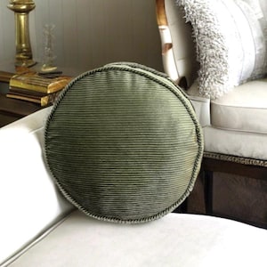 May include: A round, green, corduroy throw pillow with a decorative trim. The pillow is sitting on a white couch.