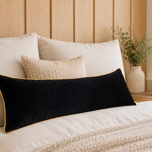 Puede incluir: Un cojín lumbar rectangular largo de terciopelo negro con un ribete amarillo brillante en los bordes. El cojín está colocado horizontalmente frente a un cojín decorativo beige texturizado y dos almohadas de cama grandes de color claro.