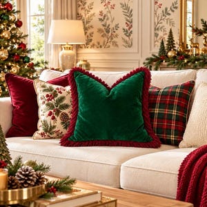 May include: A square throw pillow made of deep green velvet fabric, featuring a thick, textured dark red fringe trim along all four edges. The pillow is positioned centrally among other decorative cushions on a light-colored sofa.