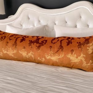 Puede incluir: Una almohada larga, rectangular, de terciopelo naranja con un patrón de damasco dorado y marrón. La almohada está sobre una cama blanca con almohadas blancas.