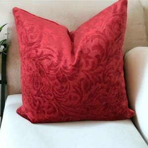 Puede incluir: Una almohada de terciopelo rojo con un patrón de damasco floral. La almohada está sentada en un sofá blanco.