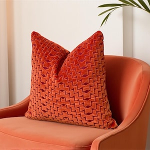 Può includere: Un cuscino decorativo arancione con un motivo geometrico poggia su una poltrona arancione abbinata. Il cuscino presenta un design a zig-zag strutturato in una tonalità più scura di arancione. La sedia ha schienale e braccioli curvi, creando un'estetica accogliente.