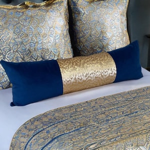 Puede incluir: Una cama con cojines decorativos. Hay dos cojines cuadrados con un diseño azul y dorado, y un cojín largo y rectangular con extremos azules y un centro dorado. También se ve un camino de cama con estampado azul y dorado.